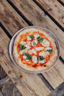 A freshly baked pizza with a thin crust, topped with tomato sauce, melted mozzarella cheese, and basil leaves is placed on a metal tray. The pizza is laid on a rustic wooden table, enhancing its artisanal and homemade appeal.