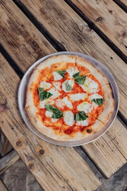A freshly baked pizza with a thin crust, topped with tomato sauce, melted mozzarella cheese, and basil leaves is placed on a metal tray. The pizza is laid on a rustic wooden table, enhancing its artisanal and homemade appeal.