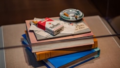 A neat stack of books sits on a table. Resting on top of the books is a red and white cigarette pack, some letters, and a booklet. An ashtray with cigarette butts is placed next to the stack.