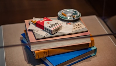 A neat stack of books sits on a table. Resting on top of the books is a red and white cigarette pack, some letters, and a booklet. An ashtray with cigarette butts is placed next to the stack.