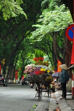 A bicycle loaded with vibrant flower bouquets stands on a sidewalk under lush green trees. The flowers include a colorful array of sunflowers, lilies, and other blooms. A person wearing a traditional conical hat is seen standing nearby, looking at the bicycle. The scene is set on a street with a few cars visible in the background, and the atmosphere is calm and serene.