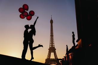 Silhouette of a couple embracing and kissing in front of the Eiffel Tower at sunset. The woman is holding red balloons, and the sky is a gradient of warm colors.