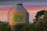 A large cylindrical metal grain silo with the words 'PINSOS JONÉ CARDEDEU' painted on it, surrounded by lush green trees and vegetation. The sky in the background features a colorful gradient from blue to pink, suggesting sunrise or sunset.