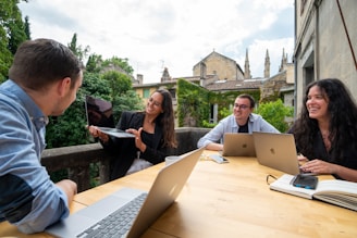 A group of four people sit around a wooden table, each with a laptop and one person holding a tablet. They are engaged in a lively discussion, smiling and appearing collaborative. The setting is outdoors on a patio with greenery and historic buildings in the background.