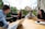 A group of four people sit around a wooden table, each with a laptop and one person holding a tablet. They are engaged in a lively discussion, smiling and appearing collaborative. The setting is outdoors on a patio with greenery and historic buildings in the background.