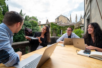 A group of four people sit around a wooden table, each with a laptop and one person holding a tablet. They are engaged in a lively discussion, smiling and appearing collaborative. The setting is outdoors on a patio with greenery and historic buildings in the background.