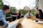 A group of four people sit around a wooden table, each with a laptop and one person holding a tablet. They are engaged in a lively discussion, smiling and appearing collaborative. The setting is outdoors on a patio with greenery and historic buildings in the background.