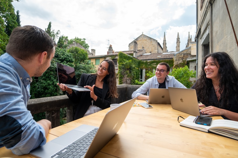 A group of four people sit around a wooden table, each with a laptop and one person holding a tablet. They are engaged in a lively discussion, smiling and appearing collaborative. The setting is outdoors on a patio with greenery and historic buildings in the background.