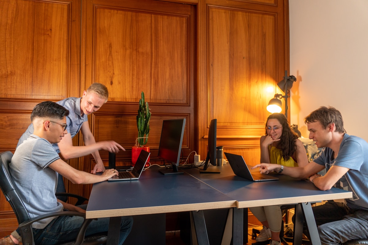 Four individuals discussing over laptops in a modern office with wooden paneling.