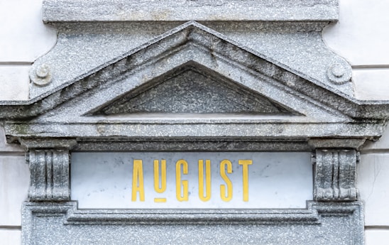 An architectural detail featuring a triangular pediment with classical design elements and the word 'August' prominently displayed in yellow letters below it.