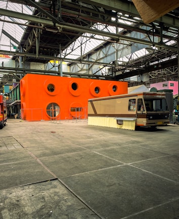 A large industrial warehouse with high ceilings and metal beams houses an orange structure with circular windows. In front of the structure is a retro-looking RV with a beige and brown color scheme. The floor is made of large concrete tiles.