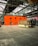 A large industrial warehouse with high ceilings and metal beams houses an orange structure with circular windows. In front of the structure is a retro-looking RV with a beige and brown color scheme. The floor is made of large concrete tiles.