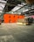 A large industrial warehouse with high ceilings and metal beams houses an orange structure with circular windows. In front of the structure is a retro-looking RV with a beige and brown color scheme. The floor is made of large concrete tiles.
