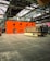 A large industrial warehouse with high ceilings and metal beams houses an orange structure with circular windows. In front of the structure is a retro-looking RV with a beige and brown color scheme. The floor is made of large concrete tiles.