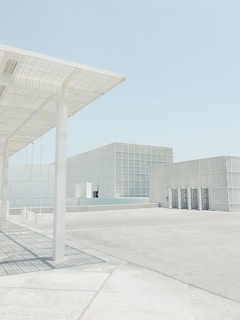A minimalist architectural scene featuring a large open space with pale concrete surfaces. Structures with a grid-like facade and a modern design are present. Soft shadows are cast by the structural elements under a clear, pale blue sky.