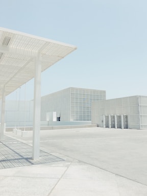 A minimalist architectural scene featuring a large open space with pale concrete surfaces. Structures with a grid-like facade and a modern design are present. Soft shadows are cast by the structural elements under a clear, pale blue sky.