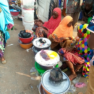 A group of people, primarily women wearing colorful traditional clothing, are seated on the ground surrounded by large bowls and buckets. Some bowls contain various items, possibly for sale, on a market street.
