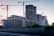 A construction site with several towering cranes and partially built high-rise buildings. The structures are primarily made of concrete and steel, and the area is surrounded by a fence with greenery in the foreground. The sky is a mix of pastel colors, suggesting sunrise or sunset, creating a serene backdrop for the urban landscape.