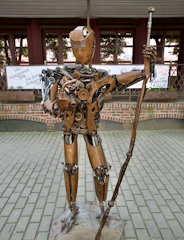 A humanoid sculpture made of mechanical and metal parts stands on a paved area. The figure includes gears, springs, and various metal components, giving it a steampunk aesthetic. It holds a rod or staff in one hand, and its face is decorated with simple facial features.