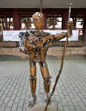 A humanoid sculpture made of mechanical and metal parts stands on a paved area. The figure includes gears, springs, and various metal components, giving it a steampunk aesthetic. It holds a rod or staff in one hand, and its face is decorated with simple facial features.
