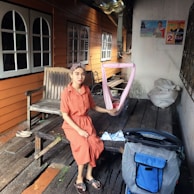 A woman in an orange dress is sitting on a rustic wooden platform, holding a pink sling with a baby inside. The background features a wooden building with several windows. Nearby, there is a wooden bench, a stroller, and some bags with miscellaneous items. Posters are visible on the white wall.