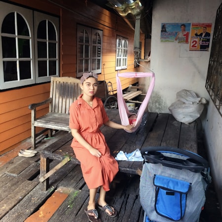 A woman in an orange dress is sitting on a rustic wooden platform, holding a pink sling with a baby inside. The background features a wooden building with several windows. Nearby, there is a wooden bench, a stroller, and some bags with miscellaneous items. Posters are visible on the white wall.