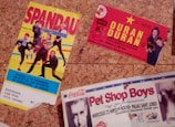 The image depicts three vintage concert tickets pinned to a corkboard. The first ticket features colorful graphics of a band called Spandau Ballet, with a predominantly yellow background. The second ticket is for a Duran Duran concert, marked by bold yellow text on a red background, with band member images in the corner. The third ticket, partially visible at the bottom, is for a Pet Shop Boys performance, showcasing an image of one of the members, along with text in red and black on a white background.
