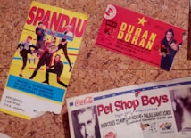 The image depicts three vintage concert tickets pinned to a corkboard. The first ticket features colorful graphics of a band called Spandau Ballet, with a predominantly yellow background. The second ticket is for a Duran Duran concert, marked by bold yellow text on a red background, with band member images in the corner. The third ticket, partially visible at the bottom, is for a Pet Shop Boys performance, showcasing an image of one of the members, along with text in red and black on a white background.