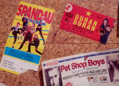 The image depicts three vintage concert tickets pinned to a corkboard. The first ticket features colorful graphics of a band called Spandau Ballet, with a predominantly yellow background. The second ticket is for a Duran Duran concert, marked by bold yellow text on a red background, with band member images in the corner. The third ticket, partially visible at the bottom, is for a Pet Shop Boys performance, showcasing an image of one of the members, along with text in red and black on a white background.