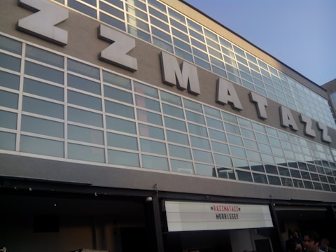 A building facade features large glass windows framed in white with the name 'Razzmatazz' displayed in bold, large letters on the exterior. Below, a marquee sign indicates an event, listing 'Razzmatazz' and 'Morrissey'. The structure seems modern with a mix of glass and concrete. People are visible at the bottom of the image, suggesting a venue entrance.
