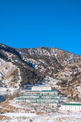 A series of buildings with green roofs are nestled at the base of a mountainous landscape. The mountain is partially covered with patches of snow and scattered trees. The sky above is clear and bright blue.