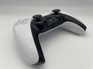 A modern video game controller with a sleek design featuring a combination of black and white colors. Two analog sticks, a directional pad, and various buttons are visible, positioned on a clean, minimalistic surface. The controller is angled slightly, giving a dynamic perspective.