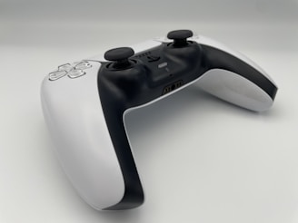 A modern video game controller with a sleek design featuring a combination of black and white colors. Two analog sticks, a directional pad, and various buttons are visible, positioned on a clean, minimalistic surface. The controller is angled slightly, giving a dynamic perspective.