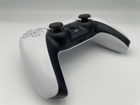 A modern video game controller with a sleek design featuring a combination of black and white colors. Two analog sticks, a directional pad, and various buttons are visible, positioned on a clean, minimalistic surface. The controller is angled slightly, giving a dynamic perspective.