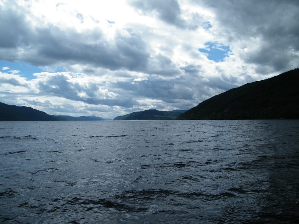 body of water near mountain under white clouds during daytime