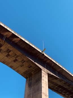 A large concrete bridge structure extends diagonally across the image against a clear blue sky. The underside of the bridge is visible, showcasing its architectural features and support pillars. One street lamp is visible on the bridge, casting a shadow on the concrete surface.