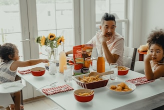 A family meal setting centered around a dining table with a variety of food items including fries and chicken nuggets. Two young children and an adult sit around the table, which is decorated with sunflowers and drinks in glass bottles. The room is bright, with natural light coming through large windows, creating a warm and inviting atmosphere.