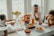 A family meal setting centered around a dining table with a variety of food items including fries and chicken nuggets. Two young children and an adult sit around the table, which is decorated with sunflowers and drinks in glass bottles. The room is bright, with natural light coming through large windows, creating a warm and inviting atmosphere.