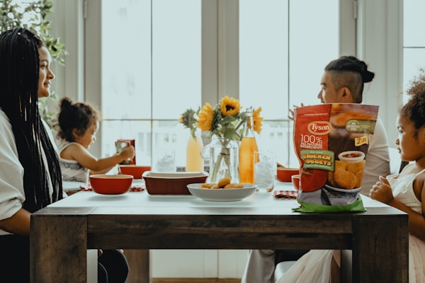 A family of four is seated around a dining table, interacting with each other. On the table, there are flower vases with sunflowers, a bowl of chicken nuggets, bottles, and cups. A bag of Tyson chicken nuggets is prominently placed on the table. Light is coming through the large windows behind the family, giving the scene a warm and bright atmosphere.