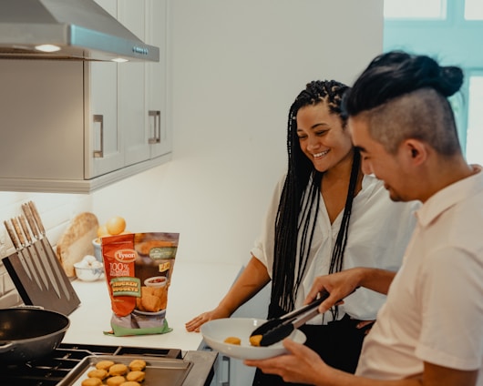 A man and woman are in a modern kitchen, smiling and preparing chicken nuggets. The man uses tongs to transfer nuggets from a baking tray to a plate. The kitchen has sleek cabinets and a set of knives on the counter. A package of Tyson chicken nuggets is prominently placed near the cooking area.