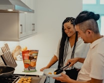 A man and woman are in a modern kitchen, smiling and preparing chicken nuggets. The man uses tongs to transfer nuggets from a baking tray to a plate. The kitchen has sleek cabinets and a set of knives on the counter. A package of Tyson chicken nuggets is prominently placed near the cooking area.