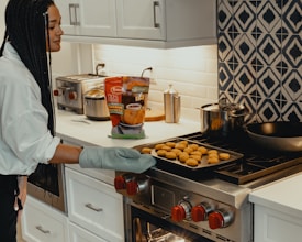 A person wearing oven mitts is removing a tray of chicken nuggets from an oven in a modern kitchen. The kitchen features white cabinets, a patterned backsplash, and various cooking appliances on the countertop.