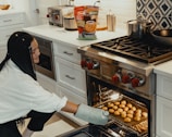 A person crouches near a modern kitchen oven, wearing an oven mitt and carefully placing a tray filled with chicken nuggets inside. The kitchen features white cabinetry, a stainless steel range, and countertop appliances including a toaster and a blender. A bag of chicken nuggets is visible on the counter.