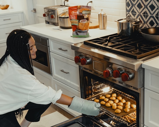 A person crouches near a modern kitchen oven, wearing an oven mitt and carefully placing a tray filled with chicken nuggets inside. The kitchen features white cabinetry, a stainless steel range, and countertop appliances including a toaster and a blender. A bag of chicken nuggets is visible on the counter.