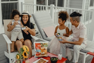 A family of four is sitting on a porch, enjoying a meal together. A woman wearing a hat is holding a child on her lap, both smiling and sharing a bowl of food. A man in a white shirt and a little girl in a white dress are seated next to them, both delighted. A table in front holds sunflowers, a packet of Tyson chicken strips, and a few bowls of food.