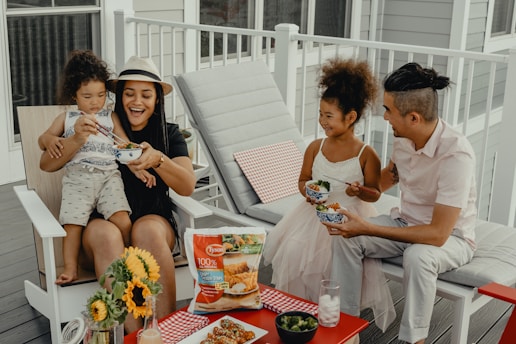 A family of four is sitting on a porch, enjoying a meal together. A woman wearing a hat is holding a child on her lap, both smiling and sharing a bowl of food. A man in a white shirt and a little girl in a white dress are seated next to them, both delighted. A table in front holds sunflowers, a packet of Tyson chicken strips, and a few bowls of food.