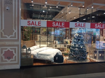 A store display showcases a cozy bedroom setup and festive decorations. A bed with white bedding is positioned beside a decorated Christmas tree with white ornaments. Surrounding furniture, including tables and chairs, adds to the homely ambiance. Red sale signs hang overhead, drawing attention to the promotional aspect.