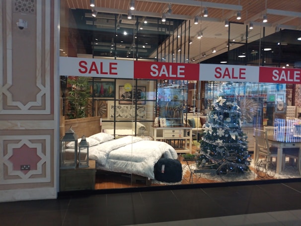 A store display showcases a cozy bedroom setup and festive decorations. A bed with white bedding is positioned beside a decorated Christmas tree with white ornaments. Surrounding furniture, including tables and chairs, adds to the homely ambiance. Red sale signs hang overhead, drawing attention to the promotional aspect.