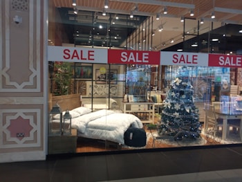 A store display showcases a cozy bedroom setup and festive decorations. A bed with white bedding is positioned beside a decorated Christmas tree with white ornaments. Surrounding furniture, including tables and chairs, adds to the homely ambiance. Red sale signs hang overhead, drawing attention to the promotional aspect.