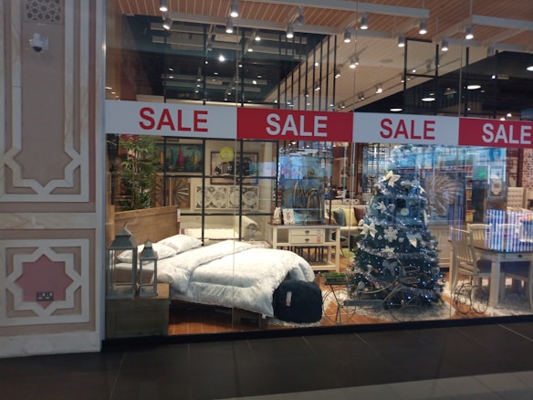 A store display showcases a cozy bedroom setup and festive decorations. A bed with white bedding is positioned beside a decorated Christmas tree with white ornaments. Surrounding furniture, including tables and chairs, adds to the homely ambiance. Red sale signs hang overhead, drawing attention to the promotional aspect.